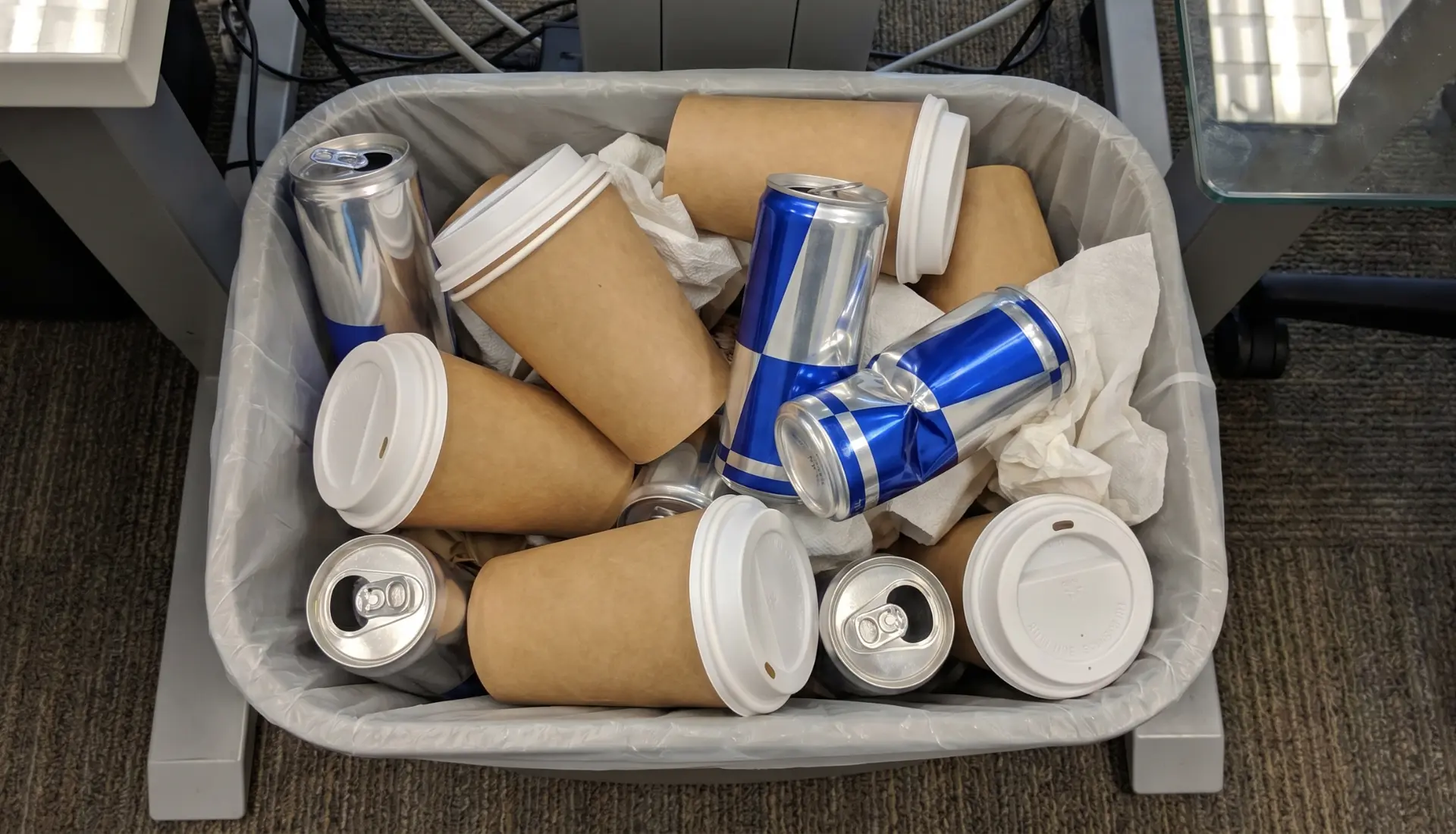 Office trash bin overflowing with empty coffee cups and crushed energy drink cans